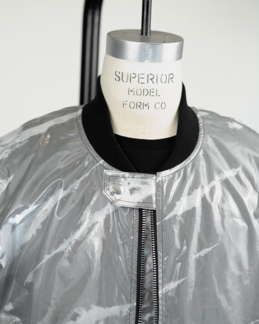 A close-up of a shiny silver JKEEFER jacket or coverall displayed on a mannequin torso. The garment has a black ribbed collar and a black front zipper, with a white mannequin neck label reading 'SUPERIOR MODEL FORM CO.