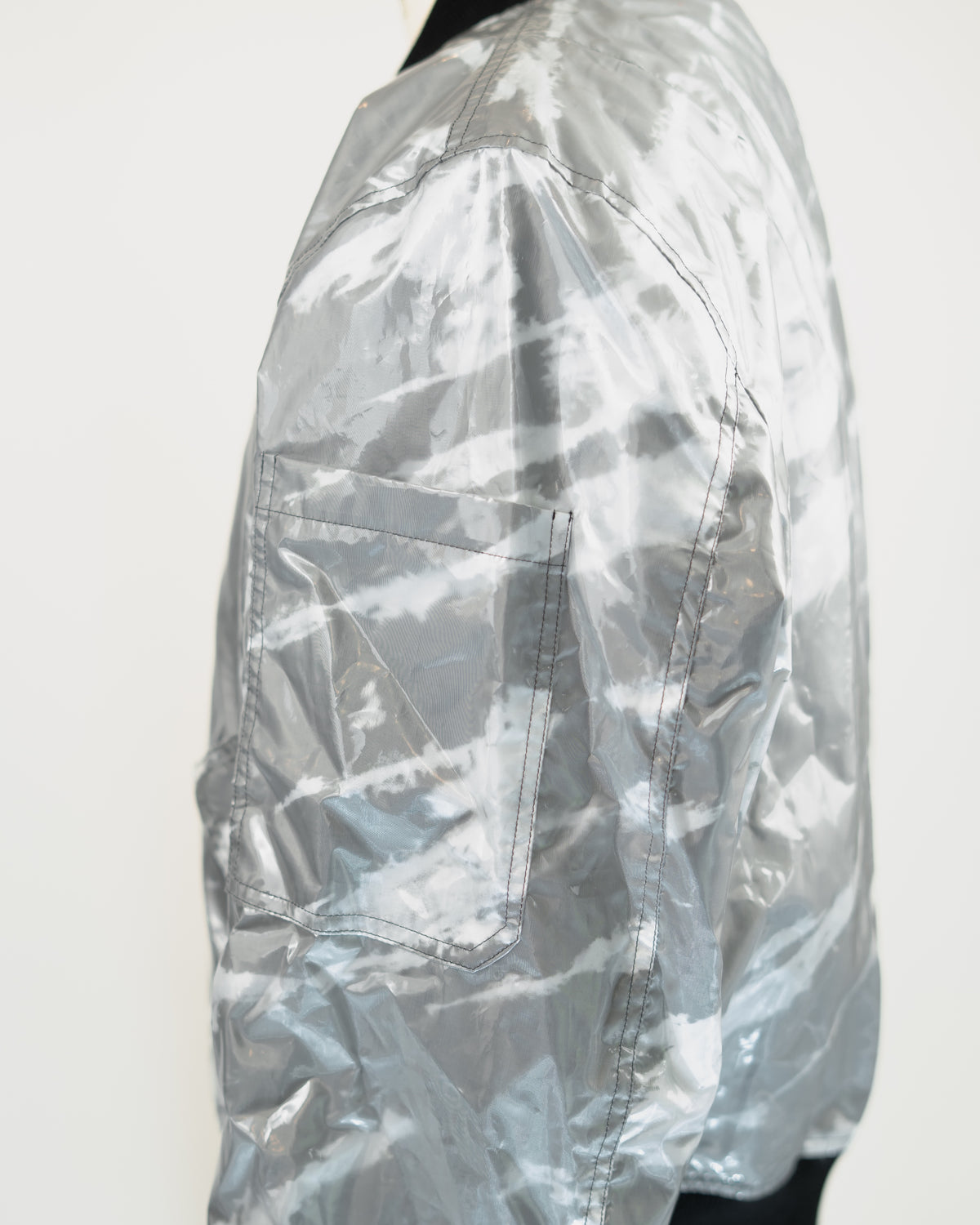 A close-up side view of a metallic silver JKEEFER jacket with a glossy, crinkled texture and a large front pocket, shown against a plain white background.