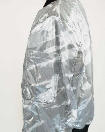 A close-up side view of a metallic silver JKEEFER jacket with a glossy, crinkled texture and a large front pocket, shown against a plain white background.