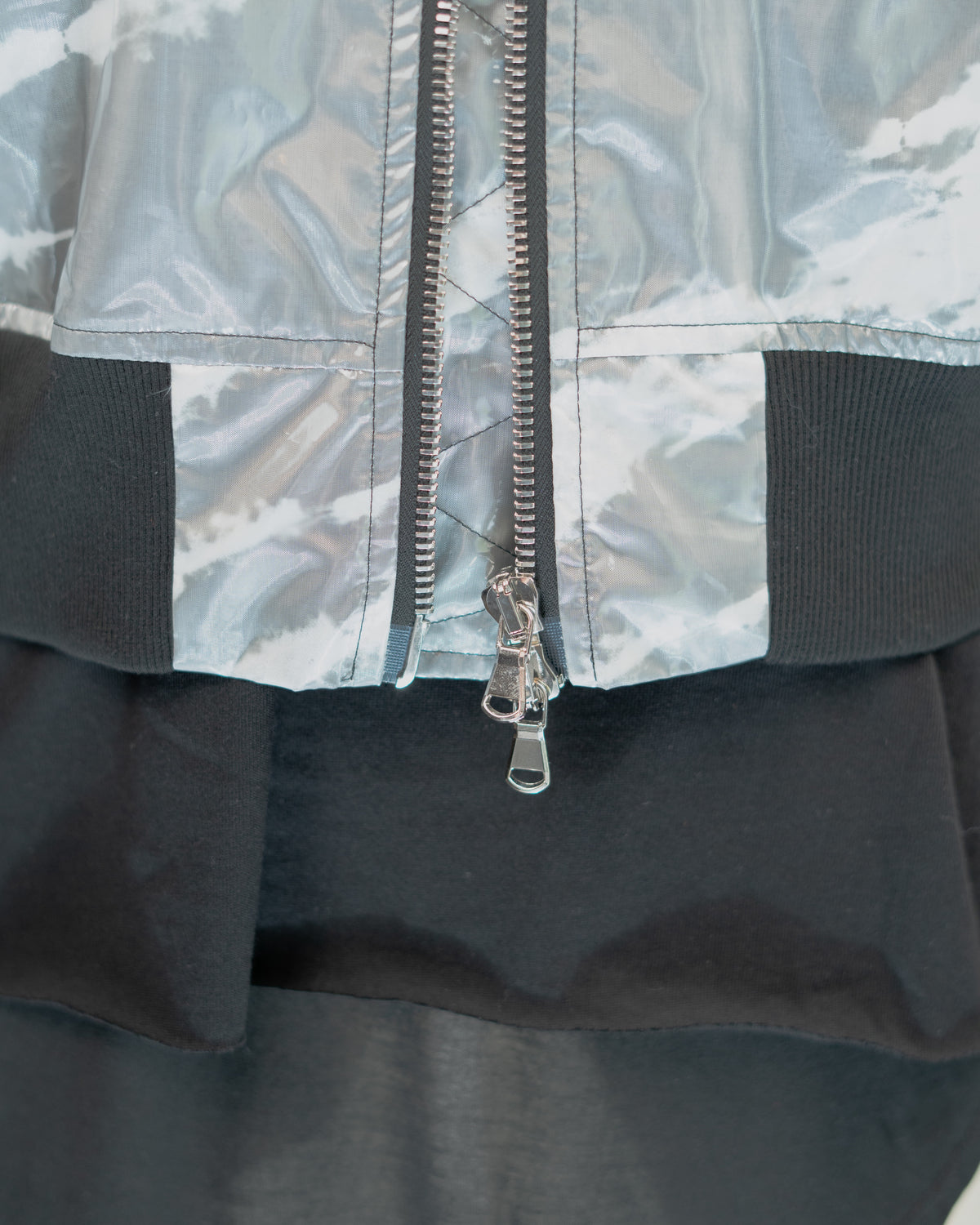 Close-up of a JKEEFER jacket detail showing a silver metallic zippered front, a light gray and white abstract patterned upper section, black ribbed trim, and a black lower section.