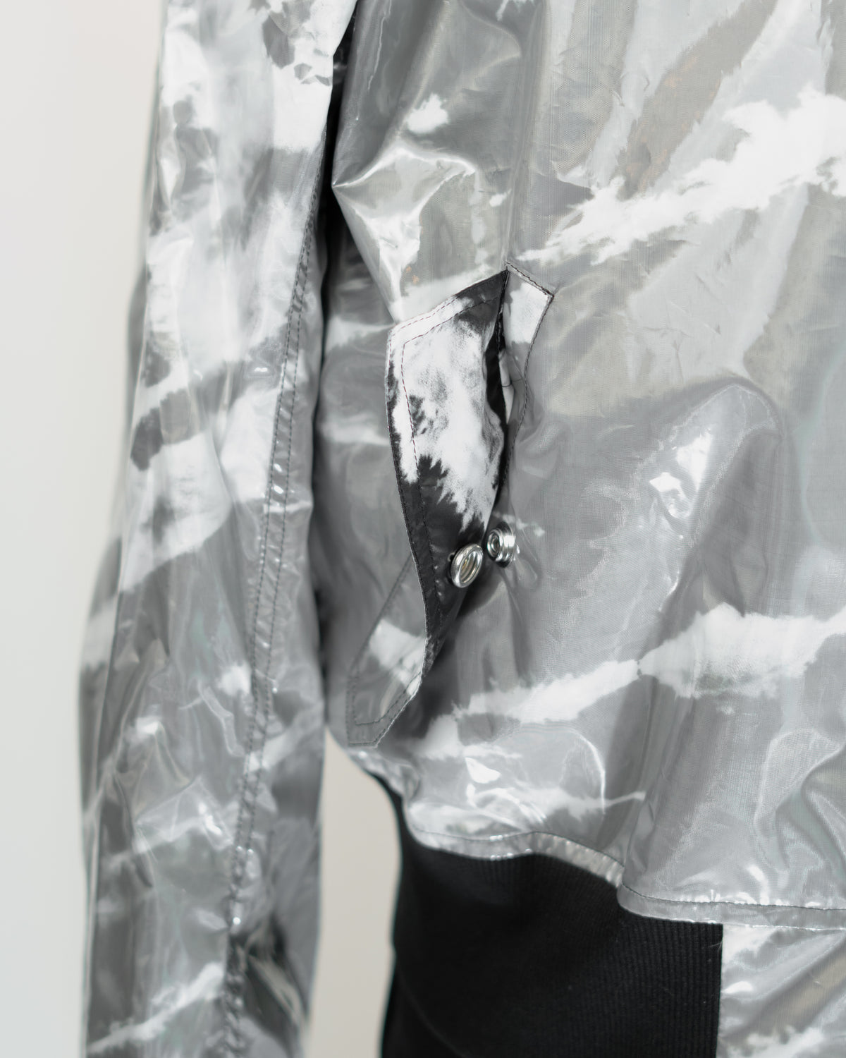 Close-up of a shiny silver-gray JKEEFER jacket with a white and gray tie-dye or marbled pattern, featuring a pocket with a black-and-white patterned flap and silver snap buttons.