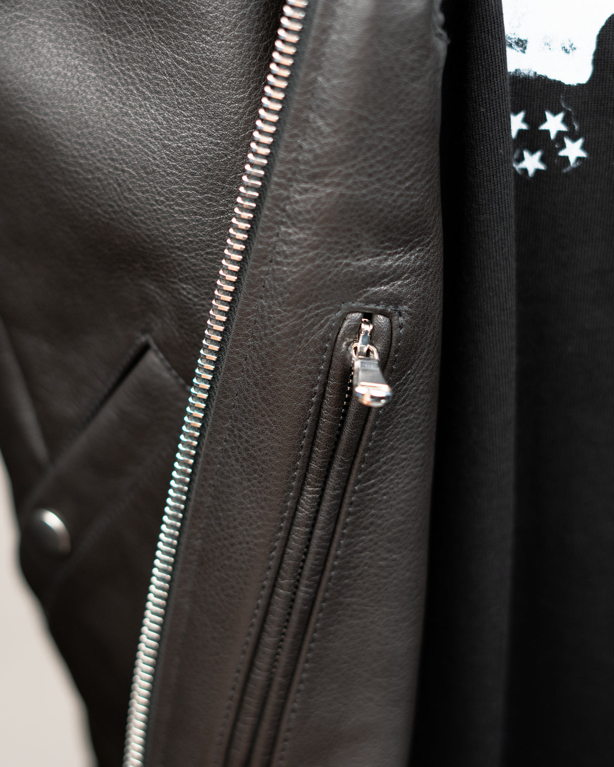 Close-up detail of a black JKEEFER leather jacket with silver metal zippers, showing the texture of the leather and a small silver zipper pull.