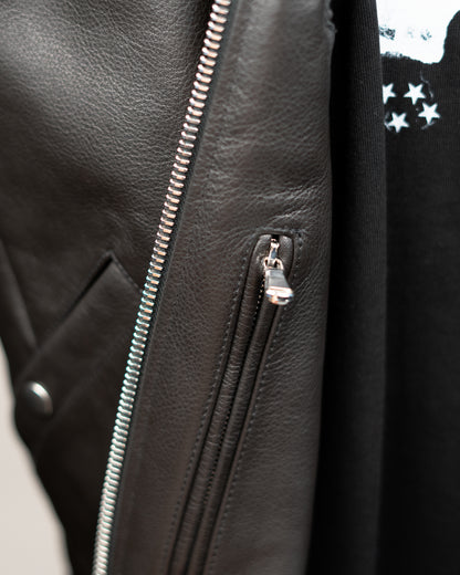 Close-up detail of a black JKEEFER leather jacket with silver metal zippers, showing the texture of the leather and a small silver zipper pull.