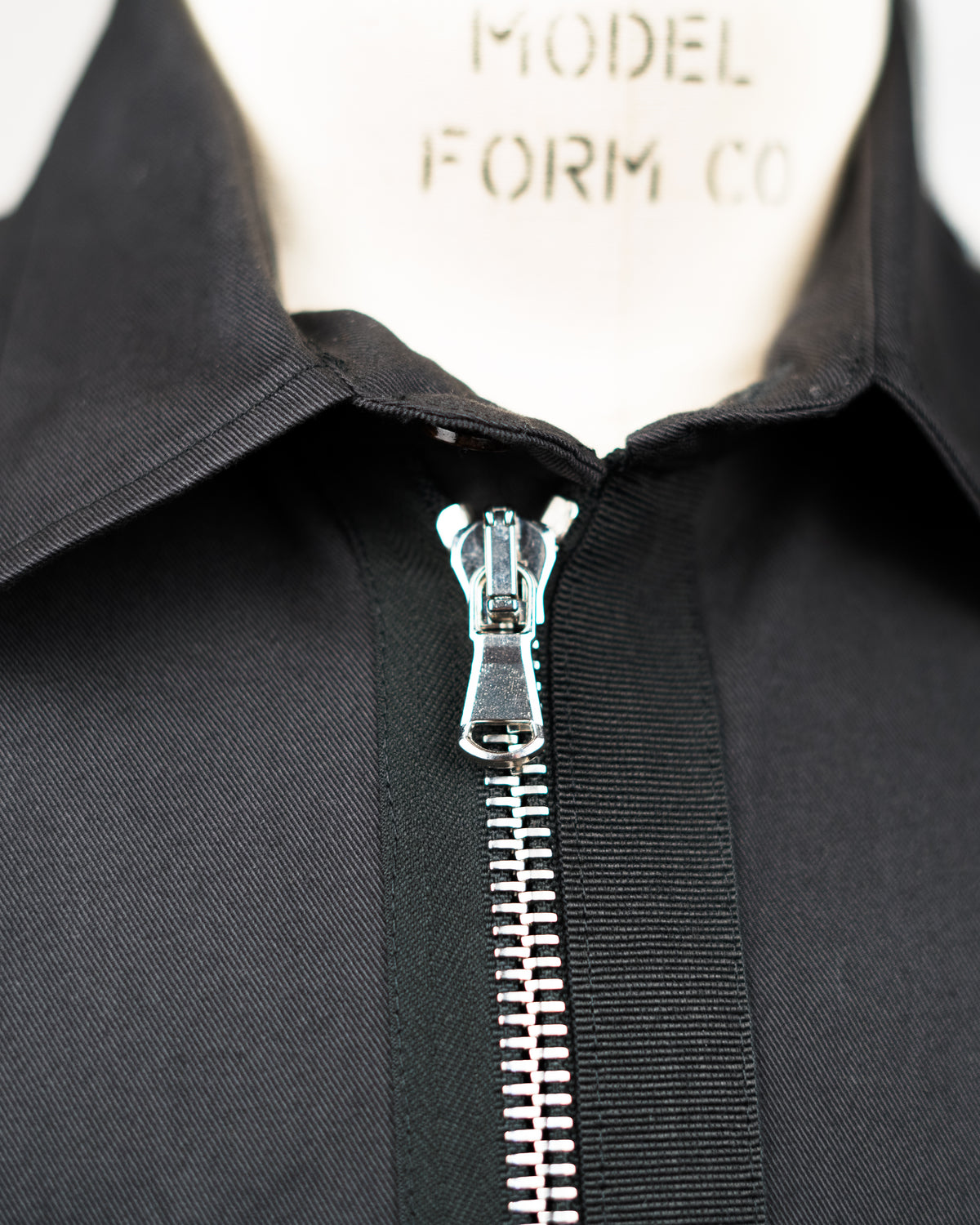 Close-up of a black JKEEFER zippered garment or jacket with a pointed collar, shown on a cream mannequin form. The zipper is open at the collar, revealing a silver metal pull and teeth.