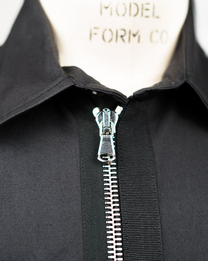 Close-up of a black JKEEFER zippered garment or jacket with a pointed collar, shown on a cream mannequin form. The zipper is open at the collar, revealing a silver metal pull and teeth.