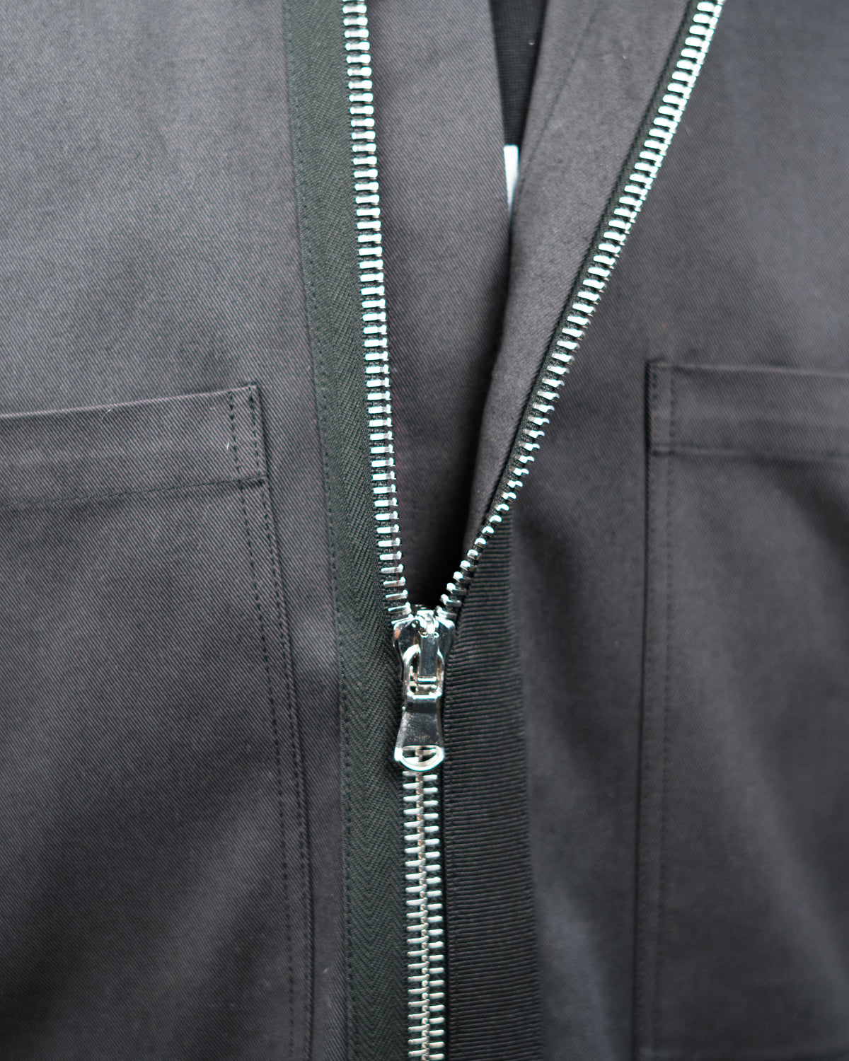Close-up of a dark gray JKEEFER zippered jacket with a silver metal zipper and a black vertical trim panel running down the center.